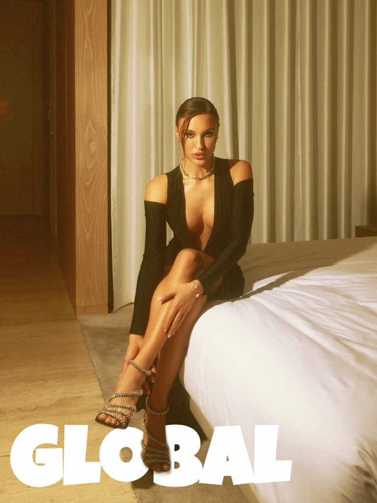 Elegant brunette escort in black evening dress sitting on hotel bed.